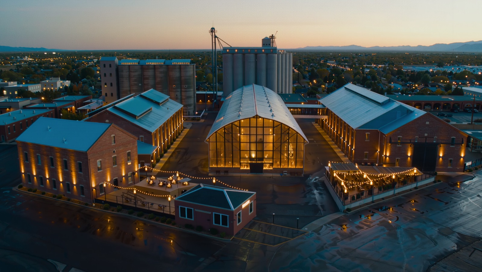 Concept render — the five-building warehouse campus at dusk, yard strung with lights, silos behind