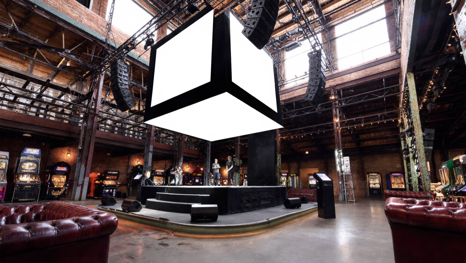 Concept render — the main hall of the five-building hub: band on the round stage beneath the hanging jumbotron cube, line-array sound rigged from the trusses, couches beside scattered machines