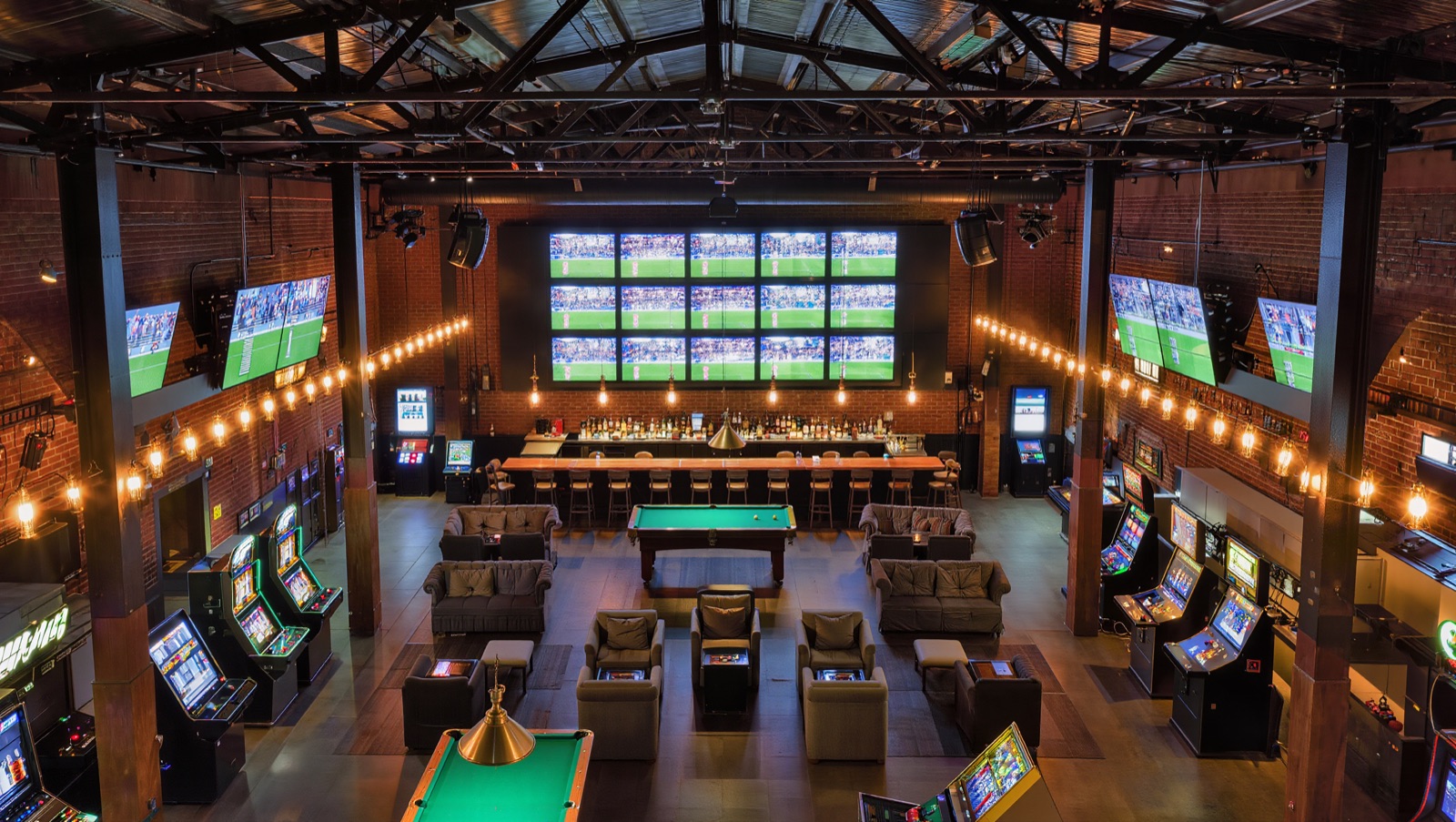 Concept render — the sports bar building: 28-foot mega-wall over the bar, pool tables, couches beside the machines
